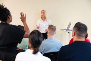 personnes de dos dont une levant la main lors d'une formation animée par une femme blonde en arrière-plan